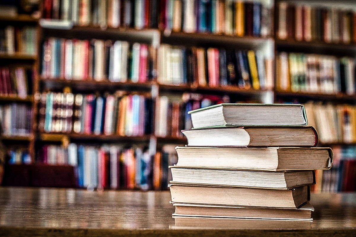 Pilha de livros sobre a mesa, estante ao fundo